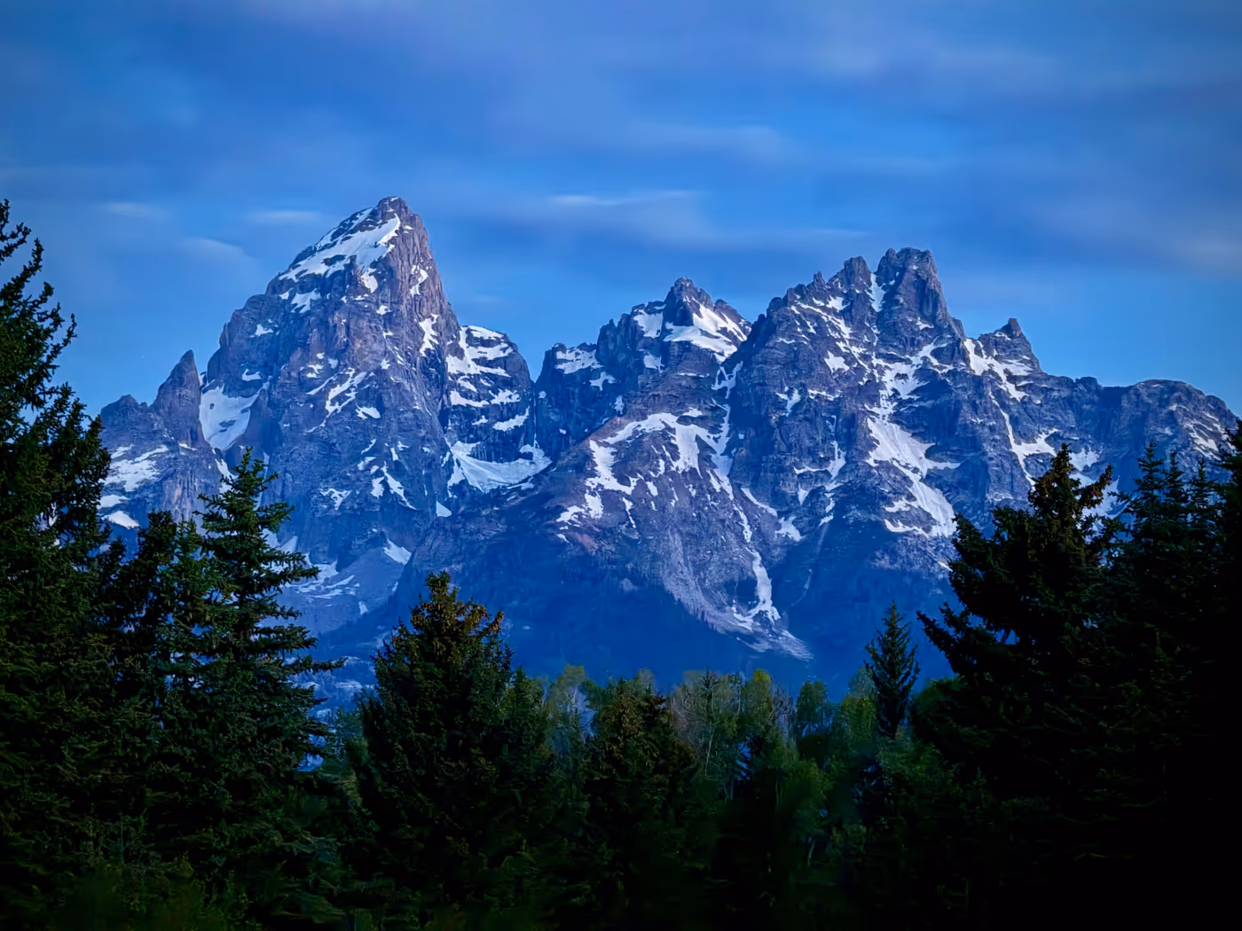 Teton peaks before sunrise in deep blue and purple light, reflected in water.