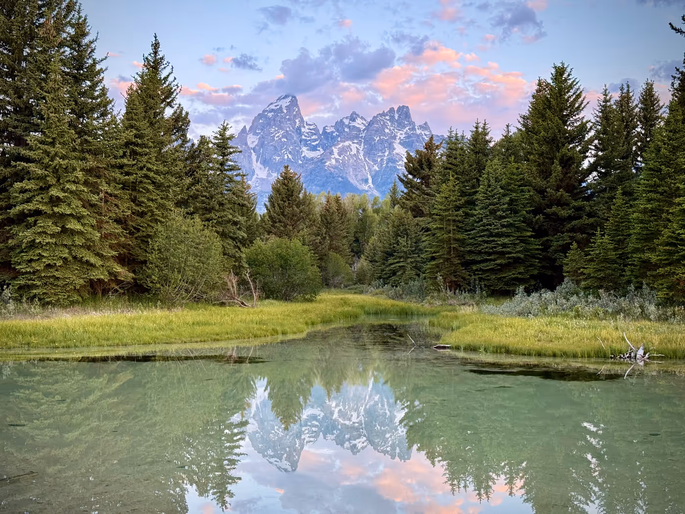 Pink dawn light on the peaks and clouds above a calm river.